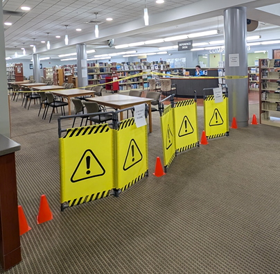 Yellow caution barriers marked with exclamation points enclosed within triangles set a perimeter in front of the reference desk. Sheets of paper taped onto the barriers read 'Wet Floor' 