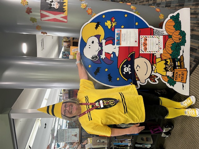 Barb, dressed up as a yellow crayon, stands beside a cardboard display featuring Peanuts characters Charile Brown, dressed as a pirate, and Snoopy, dressed as a vampire