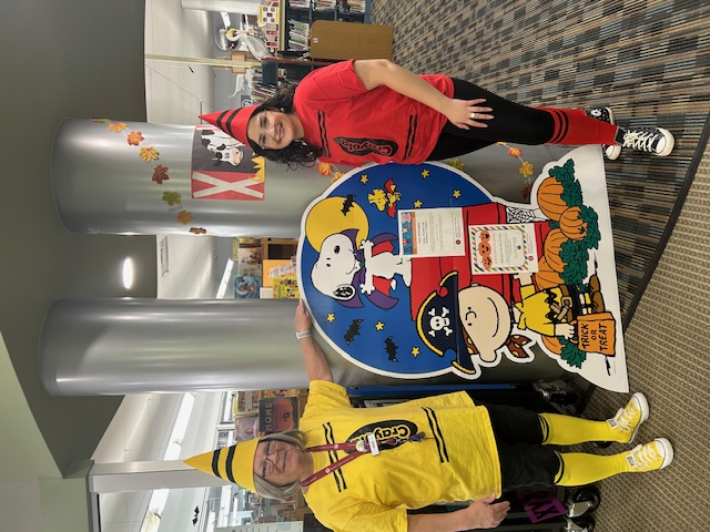 Barb, dressed up as a yellow crayon, and Rabecca, dressed up as a red crayon, stand beside a cardboard display featuring Peanuts characters Charile Brown, dressed as a pirate, and Snoopy, dressed as a vampire