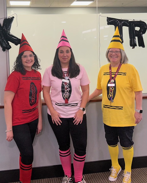 The youth services librarians dressed up as crayons. From left to right, Rabecca as red, Sara as pink, and Barb as yellow.