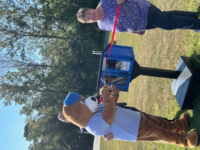 The Bells Elementary mascot, a bear in a t-shirt and cap, cuts the ribbon on the new Little Free Library