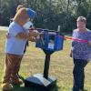 The Bells Elementary mascot, a bear in a t-shirt and cap, cuts the ribbon on the new Little Free Library